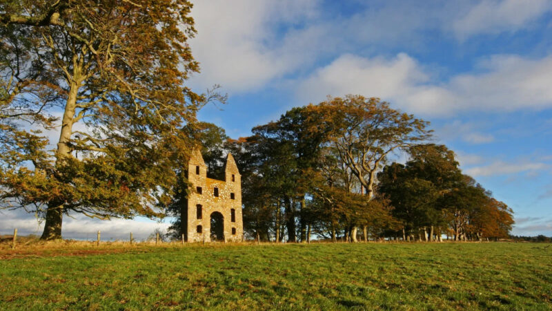 About Hundy Mundy: A tall, old stone ruin with pointed towers stands on a grassy hill, surrounded by large trees under a blue sky. Sunlight highlights the autumn foliage and the weathered façade of Hundy Mundy's impressive remains.