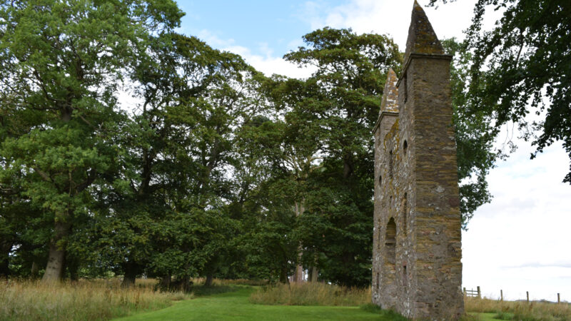 About Hundy Mundy: A stone tower ruin stands beside a grassy path, surrounded by tall green trees under a partly cloudy sky.