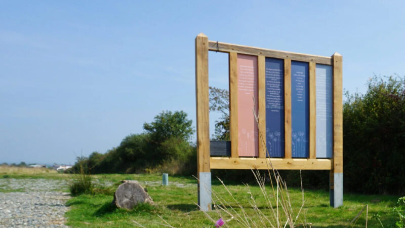 A large wooden sign with four colourful vertical panels stands on a gravel path in a natural burial meadow, surrounded by grass and bushes under a clear blue sky—a fitting tribute among memorials in Pembrokeshire.