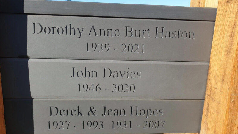 Three engraved grey stone plaques—memorials in Pembrokeshire’s natural burial meadow—bear the names and years of Dorothy Anne Burt Haston (1939–2021), John Davies (1946–2020), and Derek & Jean Hopes (1927–1993, 1931–2007).