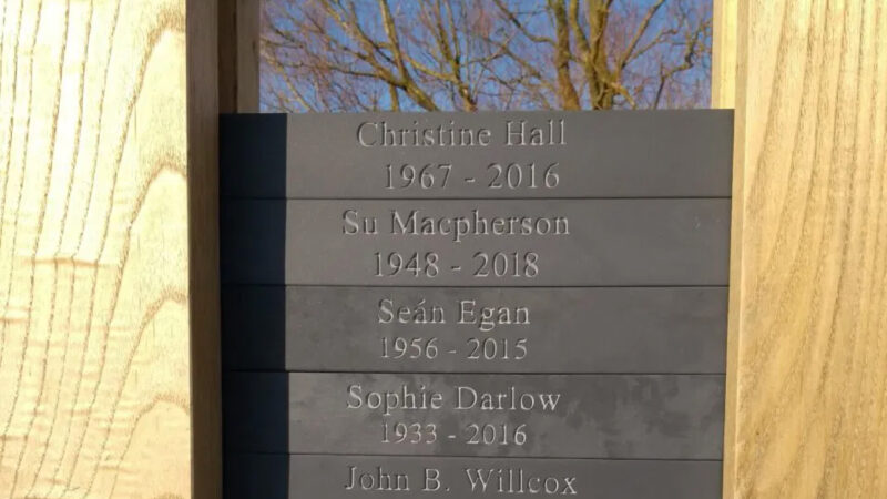 Close-up of a memorial plaque at Memorials Bath natural burial ground, engraved with names and years: Christine Hall 1967–2016, Su Macpherson 1948–2018, Seán Egan 1956–2015, Sophie Darlow 1933–2016. Trees stand in the background.