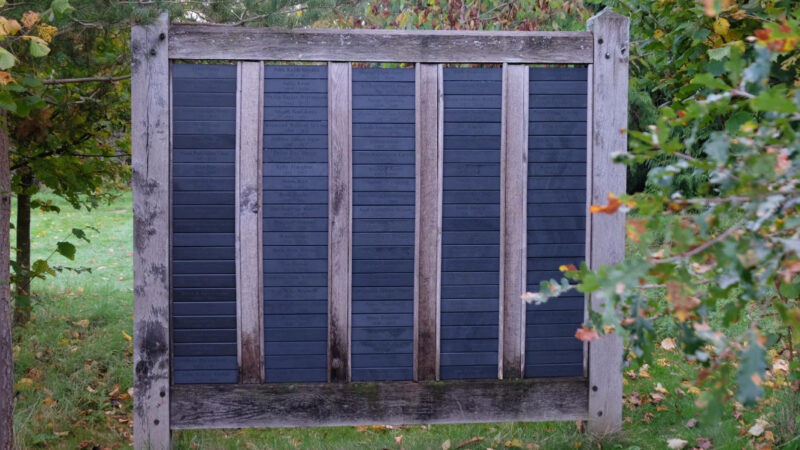 A wooden fence with evenly spaced vertical planks and a dark blue panel inset stands outdoors at Memorials Henley Woodland Burial Ground, surrounded by green grass and leafy trees with hints of autumn colours.