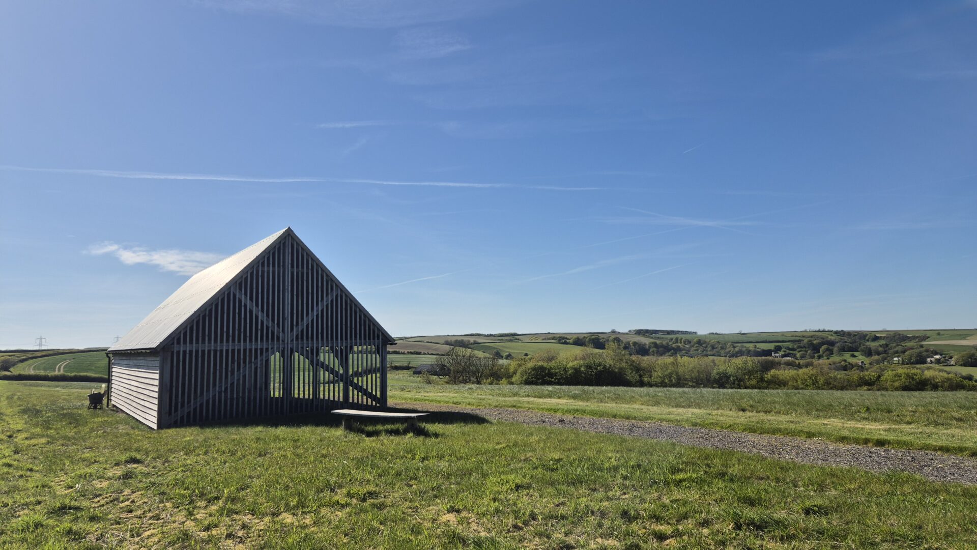 A wooden barn with a slanted roof stands on a grassy field under a clear blue sky, nestled among the rolling hills and scattered trees of the Dorset Downs, with a gravel path winding through this peaceful natural burial landscape.