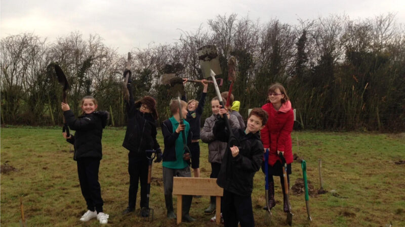 A group of children stand on grass holding spades and gardening tools, smiling at the camera near trees and bushes. They are dressed in jackets, enjoying nature close to the Aylesbury Vale natural burial memorials.