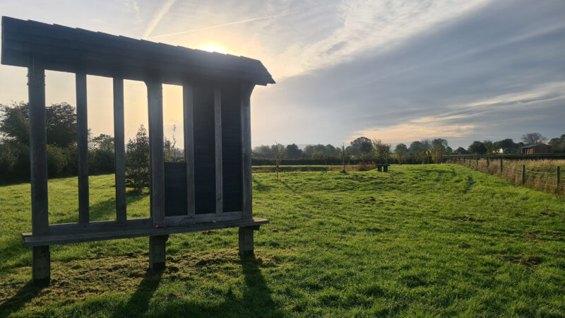 A wooden structure with vertical beams stands on a grassy field at sunrise, casting long shadows—an atmosphere reminiscent of Aylesbury Vale natural burial memorials. Trees and a fence line border the field under a partly cloudy sky.